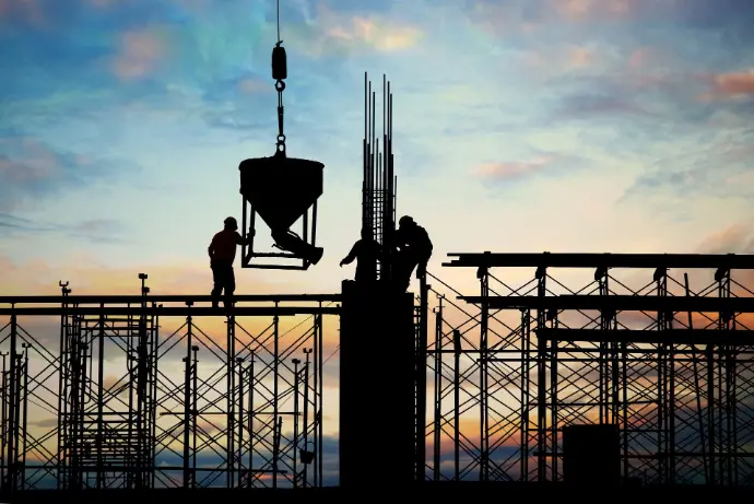 Labourers on scaffolding with a crane, and a sunset in the background.