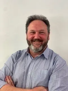 A man with a light blue button-up shirt, crossing his arms and smiling at the camera.