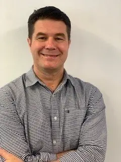 A man with a dark patterned button-up shirt, crossing his arms and smiling at the camera.